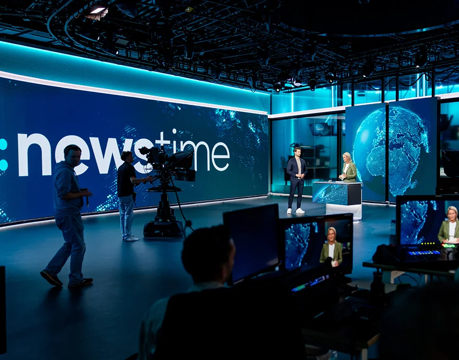 Nachrichten-Studio mit mehreren Bildschirmen und 4 Personen: jemand an der Kamera, zwei Moderatoren, eine Person gehend. Der Hintergrund zeigt groß den Schriftzug "news time". (Foto)