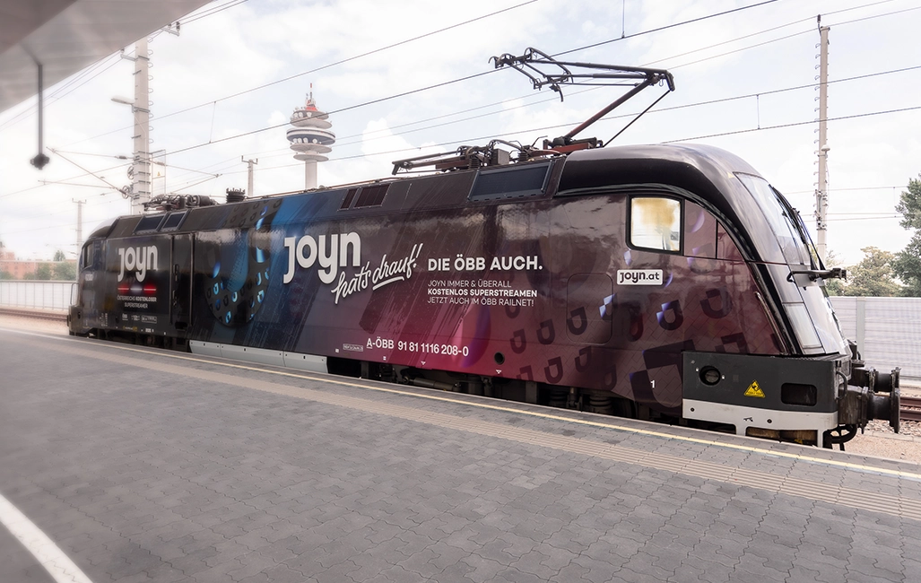 ÖBB Railjet Lok beklebt mit moderner Werbung und dem Slogan "Joyn hat’s drauf – Die ÖBB auch." Die Lok steht am Bahnsteig eines Bahnhofs. (Foto)